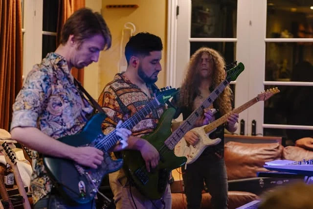 Three Jazz Odyssey musicians playing guitar and bass together in a warmly lit room.